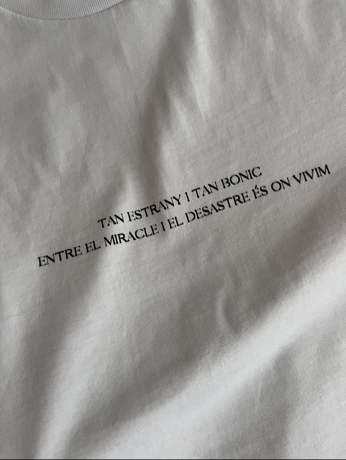 Samarreta 'Tan estrany i tan bonic entre el miracle i el desastre és on vivim' blanca amb lletres negres. Extracte de la canço 'La gent que estimo' d'Oques Grasses i Rita Payés.